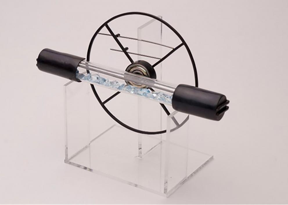 A sculptural metal picece. A clear tube filled with blue topaz is capped on either end by steel. It sits horizontally on a clear glass shelf. Behind it is a metal wheel with four spokes.